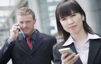 Close-up of a businesswoman holding a personal data assistant with a businessman talking on a mobile phone behind her