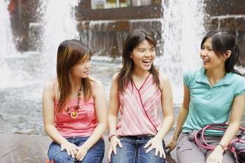Three young women sitting together at the edge of a fountain and looking cheerful