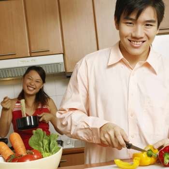Portrait of a young man cutting vegetables in the kitchen with a young woman holding noodles in the background