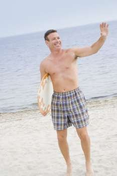 Mature man standing with a surfboard on the beach and waving his hand