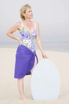 Mature woman standing on the beach with her hand on her hip and holding a body board