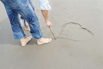 Low section view of a man standing with a woman and drawing a heart in sand with a stick