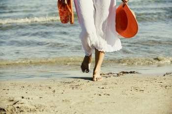Low section view of a woman holding a hat and a pair of flip-flops on the beach