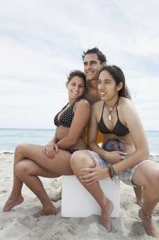 Two young women sitting on an ice box with a young man standing behind them on the beach