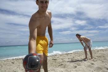 Portrait of a young man holding a soccer ball with a young woman standing behind him on the beach