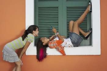 Young woman lying on a window sill and text messaging with another young woman standing beside her