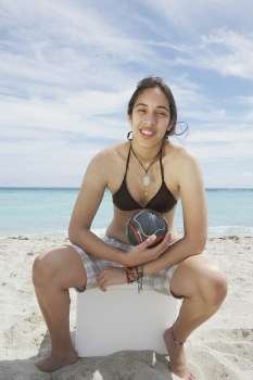 Portrait of a young woman sitting on an ice box on the beach and holding a soccer ball