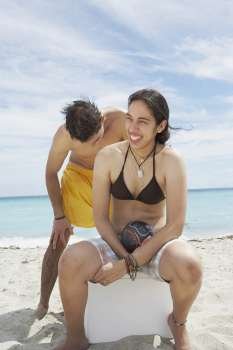 Young woman sitting on an ice box on the beach with a young man standing behind her