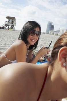 Two friends sunbathing on beach, girl on cell phone