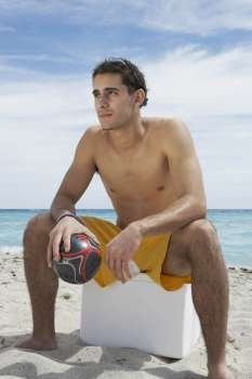 Young man sitting on an ice box on the beach and holding a soccer ball