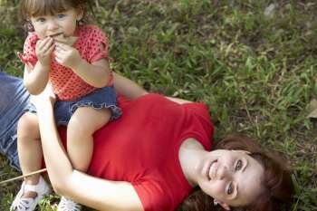 Portrait of a young woman lying on grass with her daughter sitting on her stomach