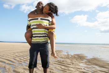 Young Man Giving Woman Piggyback Along Shoreline Of Beach