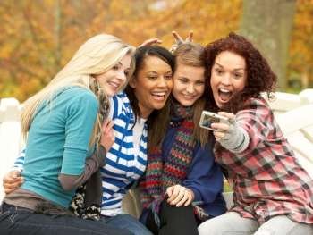Group Of Four Teenage Girls Taking Picture With Camera Sitting On Bench In Autumn Park