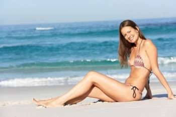 Young Woman Sitting On Sandy Beach On Holiday Wearing Bikini