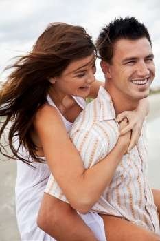 A young man and woman romantic couple having fun playing piggy back on a beach and laughing together.