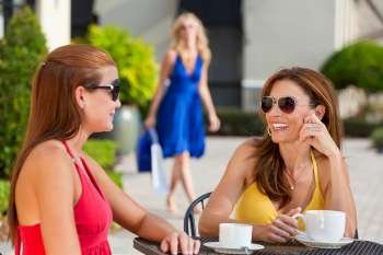 Two beautiful and sophisticated young women friends wearing sunglasses and having coffee around a modern city cafe table With their friend arriving with shopping bags in the background