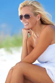A beautiful young blond woman smiling in aviator sunglasses and a white sundress sitting on a deserted tropical beach