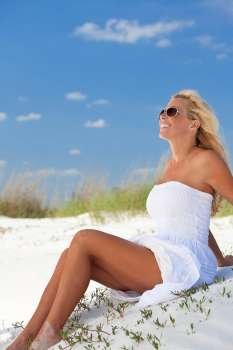 A beautiful young blond woman smiling in aviator sunglasses and a white sundress sitting on a deserted tropical beach