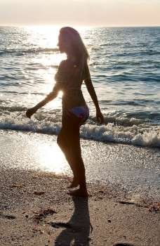 A beautiful young blond woman wearing a bikini walking along a beach at sunset
