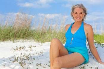 An attractive elegant senior woman in a blue swimming costume sitting on a white sand beach with grass and a blue sky behind her.