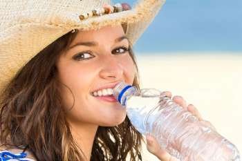 A sexy and beautiful young brunette woman sitting at the beach drinking a bottle of water with golden sand and the sea behind her