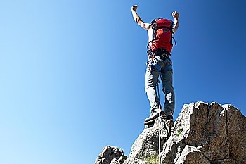 Climber reaches his arms up, standing on a stone at the top of his route, over a deep blue sky.