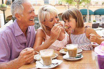 Grandparents With Granddaughter Enjoying Coffee And Cake In CafZ
