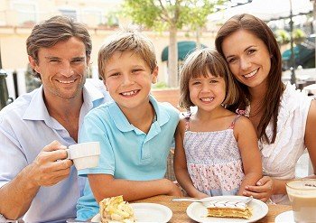 Young Family Enjoying Cup Of Coffee And Cake In CafZ Together
