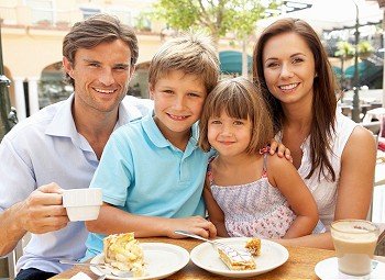 Young Family Enjoying Cup Of Coffee And Cake In CafZ Together