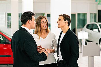 Sales situation in a car dealership, the dealer is talking to a young couple, there are cars standing in the background 