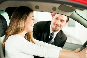 Woman buying a car in dealership sitting in her new auto, the salesman talking to her in the background