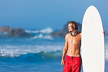 Young surfer on the beach with his surf board beside, looking at the ocean