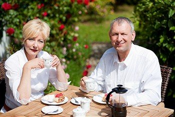 Mature or senior couple having coffee and strawberry cake on the porch in front of their home, it is summer and the roses look beautiful