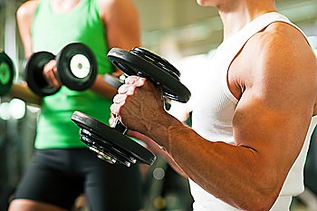 Strong man exercising with dumbbells in a gym, in the background a woman also lifting weights; focus on hands