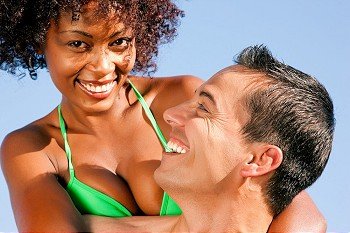 Couple in love - bikini-clad woman of color hugs a Caucasian man from behind under clear blue sky, both in beachwear in summer