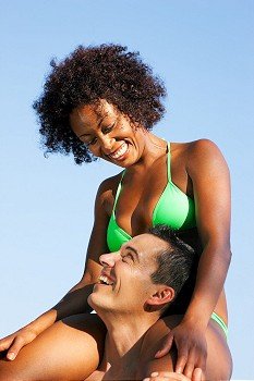 Couple in love - Woman of Brazilian origin in bikini sitting on her man’s shoulders under blue sky - summer and fun