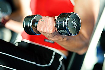 Woman lifting hand weights in a gym, in the background a man doing the same; focus on hand of woman