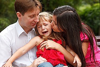 Parents calm crying girl on walk in summer garden. Girl sits in lap at father.