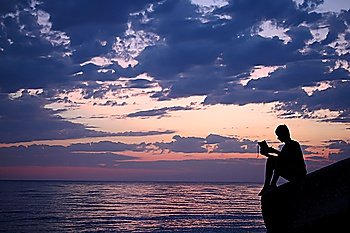 Silhouette guy sitting on breakwater in evening near sea, reads book