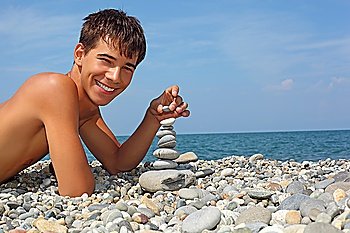 teenager boy lying on stony seacoast, creates pyramid from pebble