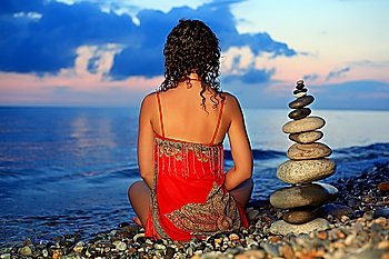 beautiful woman in red sundress sitting near to pyramid from pebble on stone seacoast in evening, sitting by back