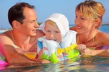 Happy family with little girl in white hat and lifejacket bathing in pool against sea, Parents looking against each other