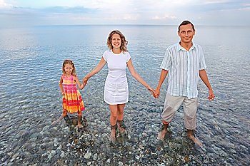 Happy family with little girl standing knee-deep in sea on beach in evening, , having joined hands