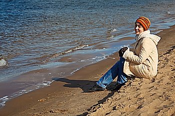 Girl sits on beach, an autumn sunny day. In cap, coat, jeans and sports shoes.