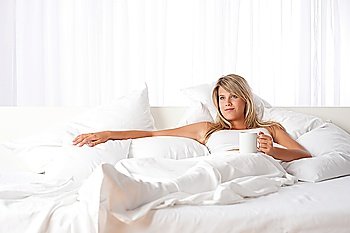 Blond woman holding cup of coffee relaxing on white bed