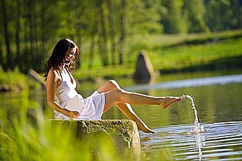 Happy romantic woman sitting by lake splashing water, wearing white dress