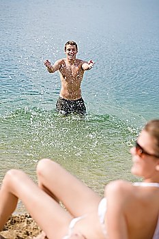 Woman in bikini sunbathing by sea on beach, focus on man splashing water