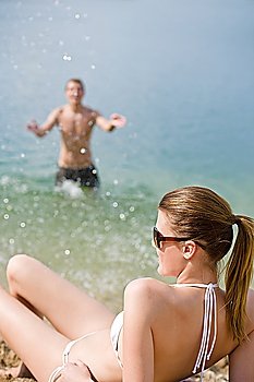 Woman in bikini sunbathing by sea on beach, man in background splashing water