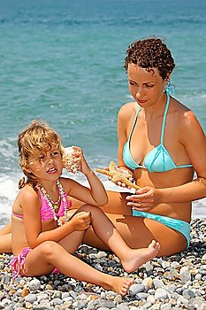 beautiful woman with pretty little girl has leant seashell bowl to an ear on stone seacoast