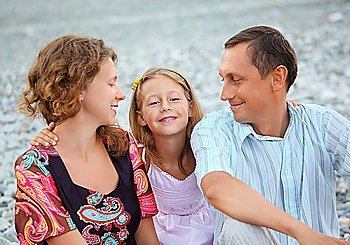 Happy family with little girl sitting on stony beach, parents looking at daughter
